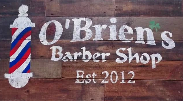 The shop’s wooden sign: O’Brien’s Barber Shop, Est 2012, painted in white on old boards beside a red, white and blue barber pole and a small green shamrock