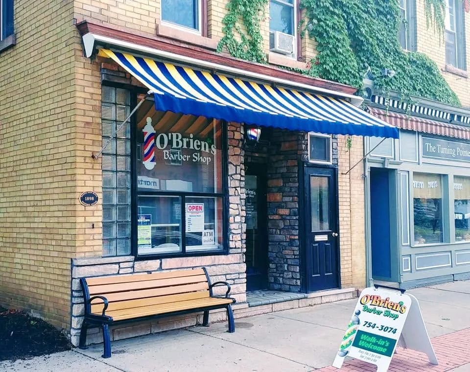 O’Brien’s Barber Shop on East Main Street in Victor: a blue and yellow striped awning, the name painted in the window beside a barber pole, a bench and a Walk-ins Welcome sign on the sidewalk